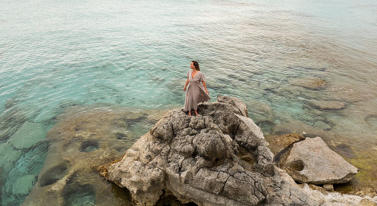 Lisette, klein in een weids kustlandschap, op een rots omringd door helder turquoise water.